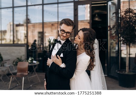 Stylish newly married European couple. Smiling bride in white dress hugging the groom. Groom with a beard and glasses dressed in a classic black suit, white shirt and bow-tie sitting. Outdoors wedding Royalty-Free Stock Photo #1775251388