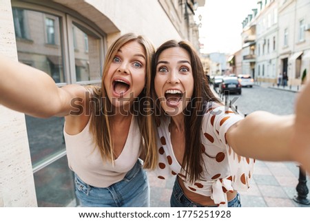 Image of excited adult two women hugging and taking selfie photo while walking on city street