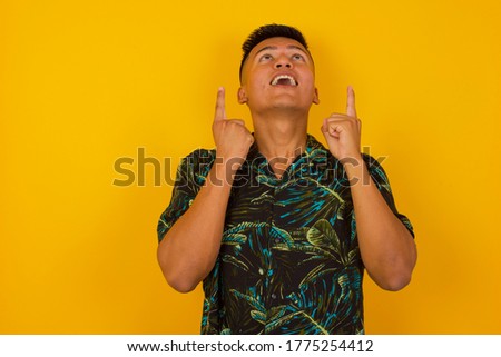 Charming carefree man with positive expression, points up with both index fingers, dressed in casual clothing, has broad intrested smile, isolated over pink background. Look there, please.