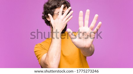 young crazy cool man covering face with hand and putting other hand up front to stop camera, refusing photos or pictures against flat wall
