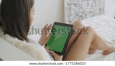 View over head on of caucasian woman lying on the bed at home. Girl holding tablet computer with green screen while watching something. Chroma key