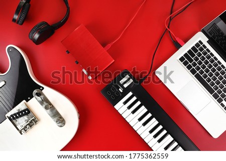 Top view of home recording studio equipment, electric guitar, MIDI keyboard, audio interface and headphone on red background.
