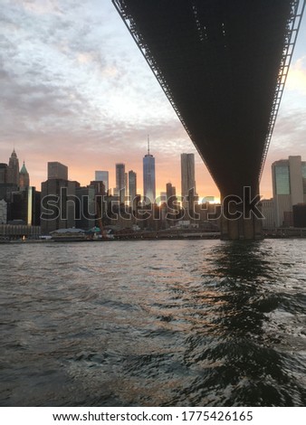 Going under the Brooklyn Bridge in a ferry on the East River, view of lower Manhattan skyline at sunset, Tall New York City skyscrapers late evening, dramatic colorful sky. Sun setting on Manhattan.