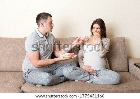 Husband offers donuts to his pregnant wife but she refuses and makes stop gesture because she feels sick. Feeling bad during pregnancy concept.