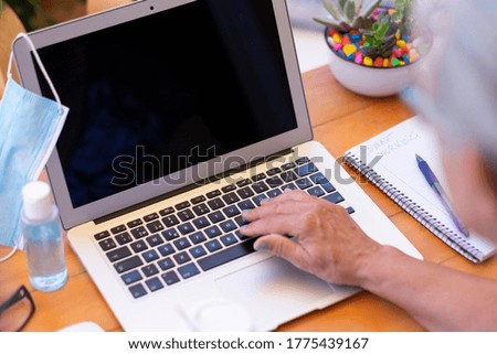 Defocused senior woman typing on laptop keyboard, mask and disinfectant on the table, the new normal at the time of the coronavirus