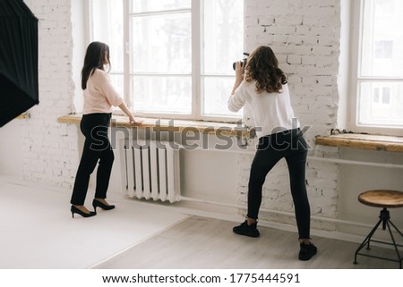 Young woman photographer shooting attractive female model on digital camera in light photo studio, on background large window. Concept of creative work in photo studio, backstage job.