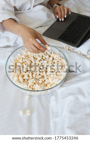 The girl is sitting on the bed with her laptop open and eating popcorn. Watching an interesting movie. Light food. Healthy lifestyle.