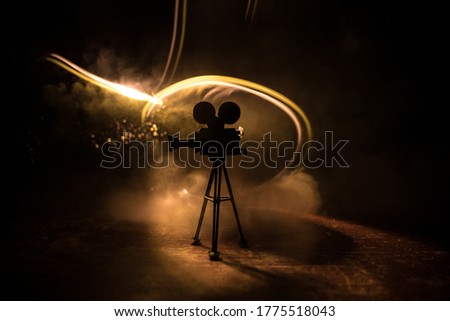 Movie concept. Miniature movie set on dark toned background with fog and empty space. Silhouette of vintage camera on tripod. Selective focus Royalty-Free Stock Photo #1775518043