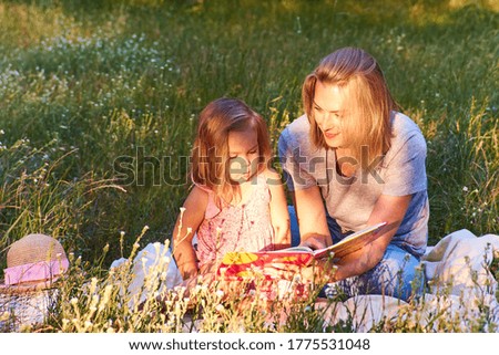 Mother and little daughter reading book sitting on green grass outdoor in park Summer time Happy family 
