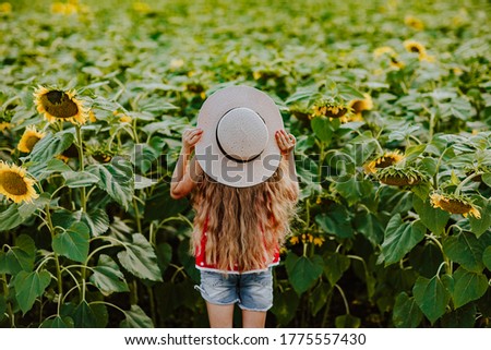 Cute young blond girl in red summer shirt and straw hat posing at the sunflower's field.