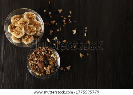 Bananas cut into slices with granola and cereal on dark gray wooden background in glass bowls on a table. Top view, copy space.