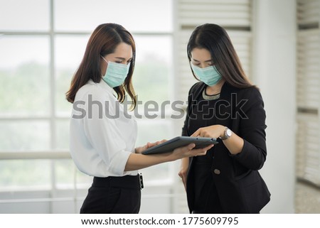 Two female office workers in business suit wears facemask and discuss about their work via tablet in business building. Businesswoman talking and poitning on tablet. Business stock photo