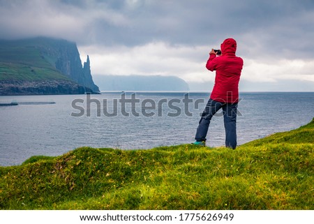 Tourist takes picture of Trollkonufingur cliffs on Vagar island. Dramatic summer scene of outskirts of Sandavagur village, Faroe Islands, Kingdom of Denmark, Europe.