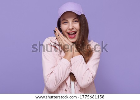 Portrait of glad full of happiness girl with wide open mouth and happy eyes, keeping palms together near cheek, looking at camera isolated on lilac background.