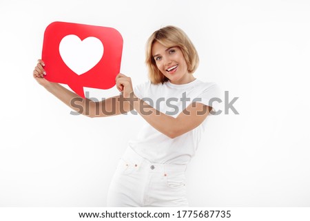 Gleeful young blonde woman holding speech bubble heart like symbol placard over red background.