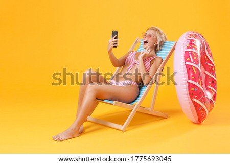 Funny young woman in pink striped swimsuit glasses sit on deck chair isolated on yellow background. People summer vacation rest lifestyle concept. Doing selfie shot on mobile phone, making video call