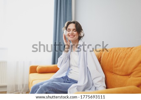 Charming young woman girl in white casual clothes sit on couch spending time in living room at home. Rest relax good mood leisure lifestyle concept. Listen music with headphones, keeping eyes closed