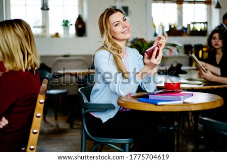 Confident young woman in elegant clothes listening to voice message on mobile phone and smiling while relaxing in contemporary cafeteria