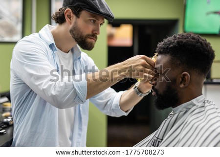 Barber trim hair with clipper on young unshaven black man in barbershop studio. Professional hairdresser cut hair with electric shearer machine on African guy.