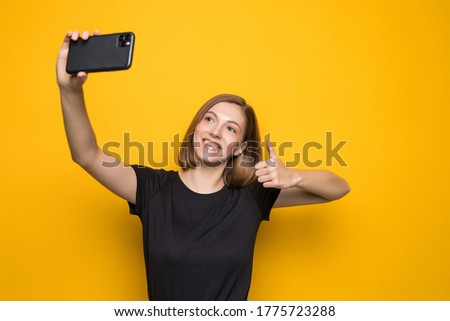 Shouting young woman taking a selfie photo on yellow background.