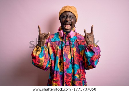 Young handsome african american man wearing colorful coat and cap over pink background shouting with crazy expression doing rock symbol with hands up. Music star. Heavy concept.
