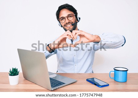 Handsome hispanic man working at the office wearing operator headset smiling in love doing heart symbol shape with hands. romantic concept. 