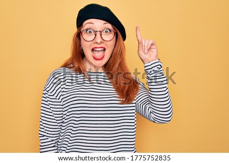 Beautiful redhead woman wearing striped t-shirt and french beret over yellow background pointing finger up with successful idea. Exited and happy. Number one.