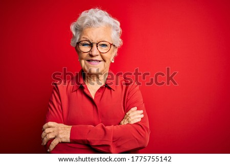 Senior beautiful grey-haired woman wearing casual shirt and glasses over red background happy face smiling with crossed arms looking at the camera. Positive person.