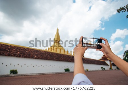 Tourist girl in Pha That Luang, taking photo of old measure with smartphone.