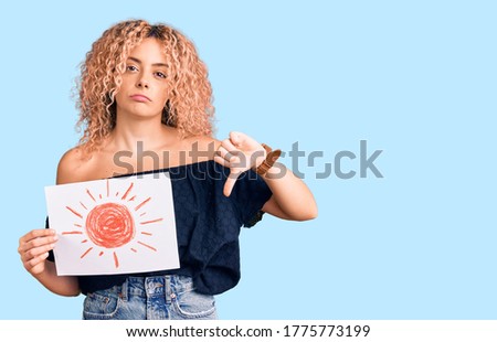 Young blonde woman with curly hair holding sun draw with angry face, negative sign showing dislike with thumbs down, rejection concept 
