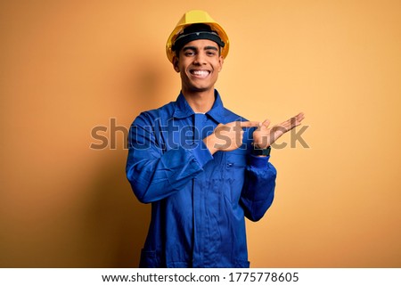 Young handsome african american worker man wearing blue uniform and security helmet amazed and smiling to the camera while presenting with hand and pointing with finger.