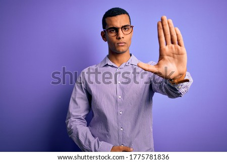 Handsome african american man wearing striped shirt and glasses over purple background doing stop sing with palm of the hand. Warning expression with negative and serious gesture on the face.