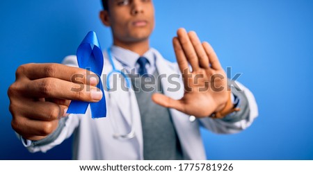 Young handsome african american doctor man holding blue cancer ribbon symbol with open hand doing stop sign with serious and confident expression, defense gesture