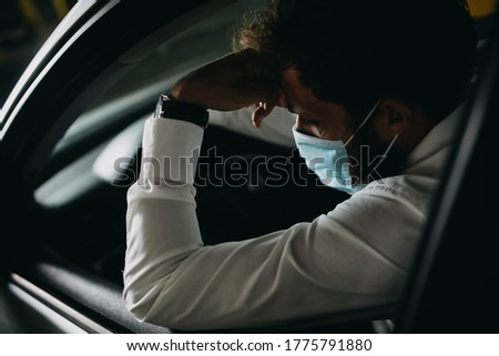 A depressed man with a protective mask on his face behind the wheel of a car. Covid-19