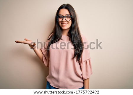 Young brunette elegant woman wearing glasses over isolated background smiling cheerful presenting and pointing with palm of hand looking at the camera.