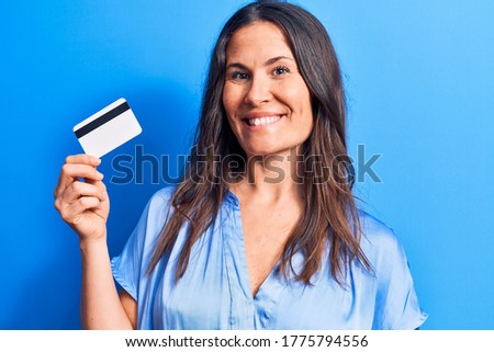 Young beautiful brunette woman holding credit card over isolated blue background looking positive and happy standing and smiling with a confident smile showing teeth