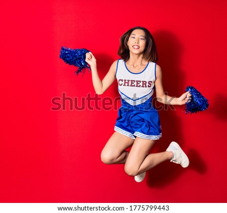 Young beautiful chinese girl smiling happy wearing cheerleader uniform. Jumping with smile on face using pompom over isolated red background