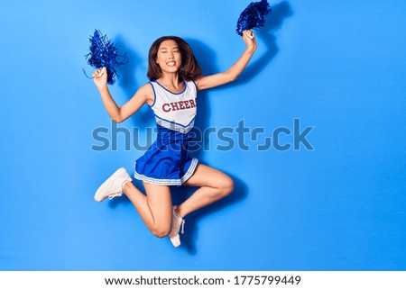 Young beautiful chinese girl smiling happy wearing cheerleader uniform. Jumping with smile on face using pompom over isolated blue background