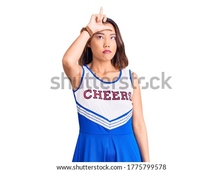 Young beautiful chinese girl wearing cheerleader uniform making fun of people with fingers on forehead doing loser gesture mocking and insulting. 