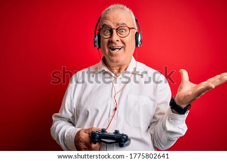 Middle age handsome hoary gamer man playing video game using joystick and headphones very happy and excited, winner expression celebrating victory screaming with big smile and raised hands