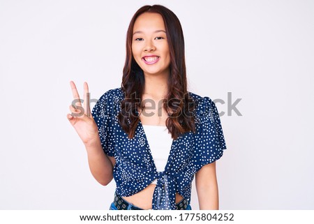 Young beautiful chinese girl wearing casual clothes smiling with happy face winking at the camera doing victory sign. number two. 