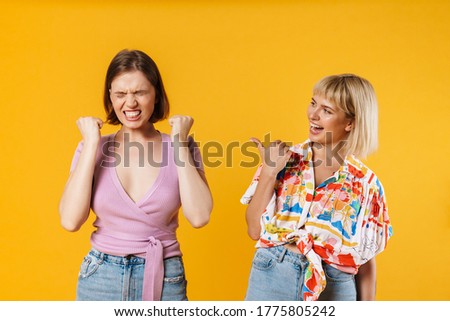 Portrait of two cheerful lovely girlfriends wearing summer clothes standing isolated over yellow background, celebrating success