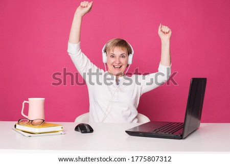 Old woman in her 60s works on computer, wearing headphohes. Laptop on white table and Pink background. She make happy winning gestures