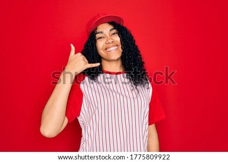 Young african american curly sportswoman wearing baseball cap and striped t-shirt smiling doing phone gesture with hand and fingers like talking on the telephone. Communicating concepts.