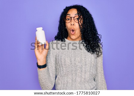 Young african american curly woman holding pills standing over isolated purple background scared in shock with a surprise face, afraid and excited with fear expression