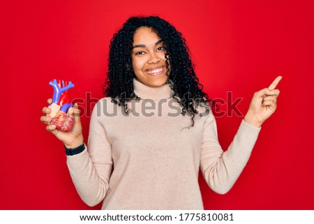 Young african american curly woman holding plastic heart standing over red background very happy pointing with hand and finger to the side