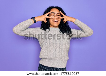Young african american woman wearing casual sweater and glasses over purple background Doing peace symbol with fingers over face, smiling cheerful showing victory