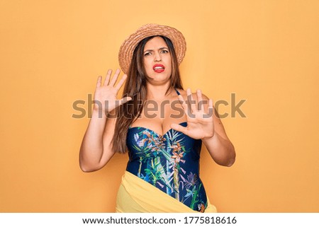 Young hispanic woman wearing summer hat and swimsuit over yellow background afraid and terrified with fear expression stop gesture with hands, shouting in shock. Panic concept.