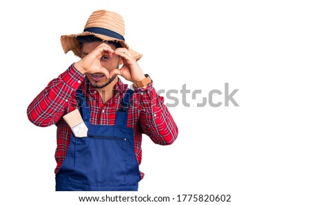 Handsome latin american young man weaing handyman uniform doing heart shape with hand and fingers smiling looking through sign 
