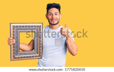 Handsome latin american young man holding empty frame smiling happy and positive, thumb up doing excellent and approval sign 